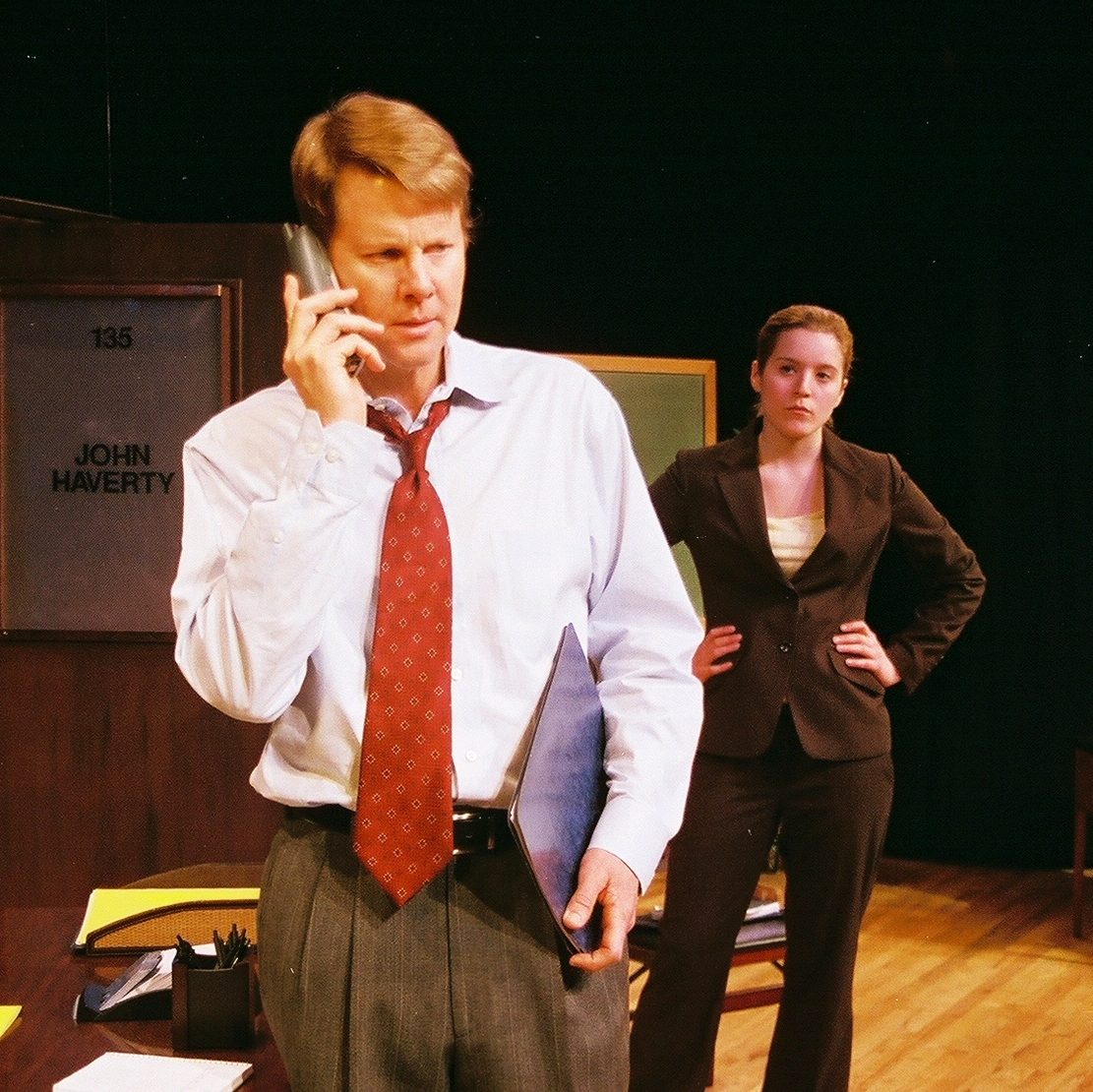 A man with a concerned expression speaks on a phone in the foreground while a woman stands behind him with her hands on her hips. The photo is from a 2006 production of Oleanna by David Mamet.